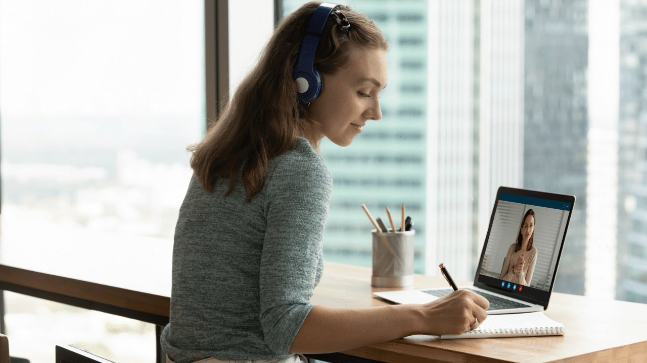 woman sitting at desk with headphones, taking notes as she participates on Zoom call